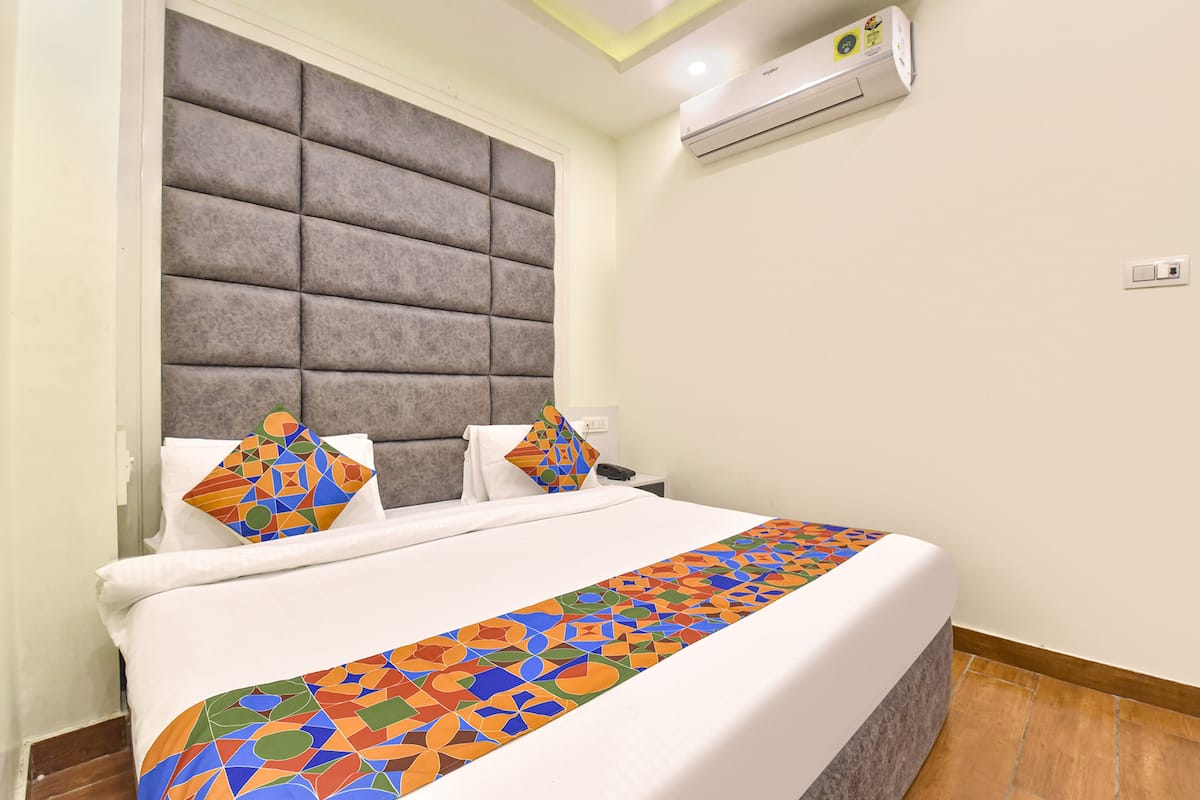 A hotel room with a large bed, a grey upholstered headboard, a patterned bedspread, and an air conditioning unit on the wall.