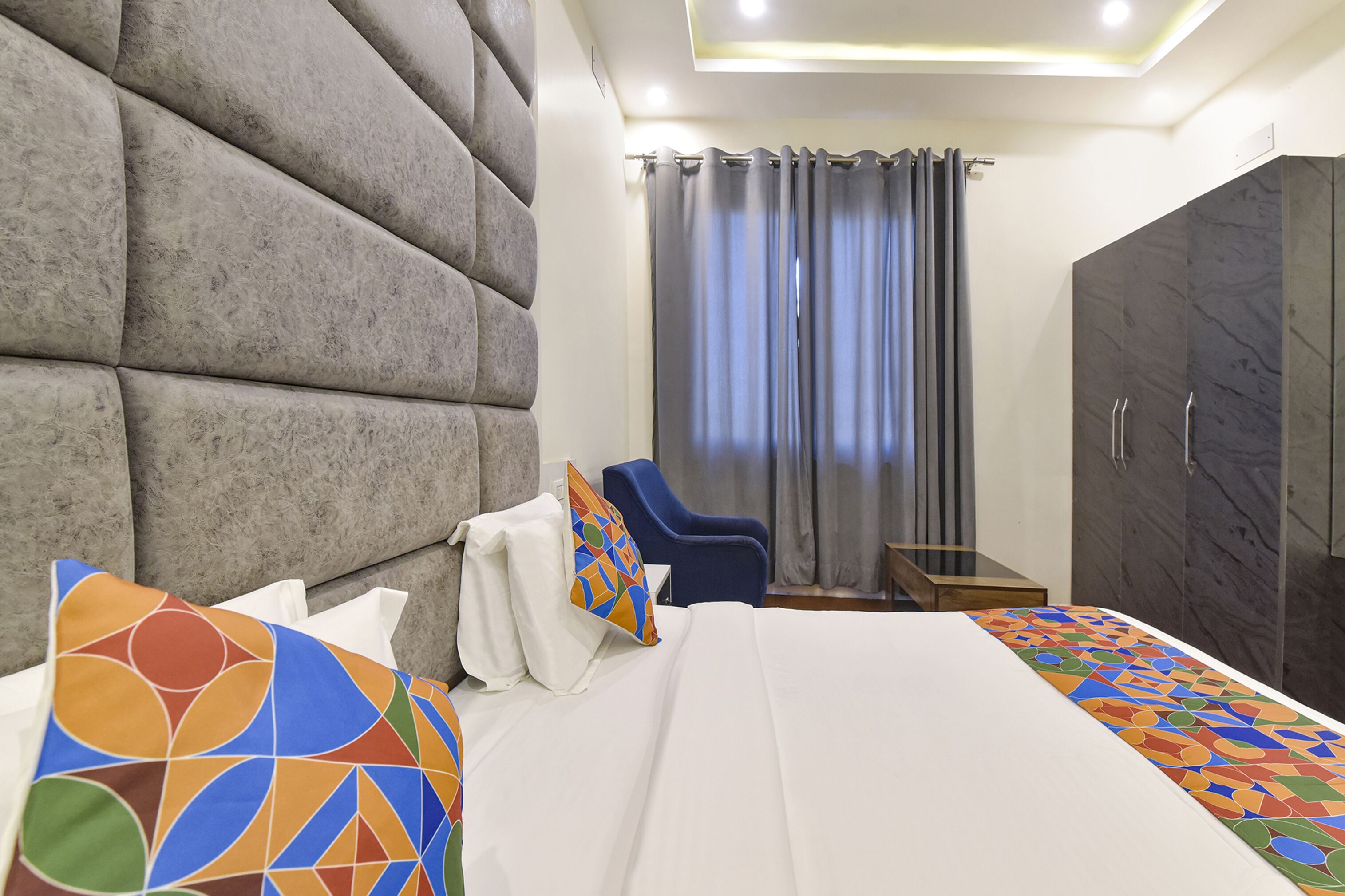 A modern hotel room with a large bed, a seating area with a chair, a wardrobe, and a view of the outside through curtains.