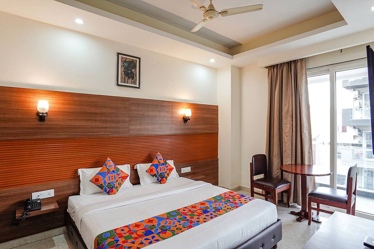 Habitación de hotel con una cama grande, cabecera de madera, dos lámparas de pared, una mesita, una silla y un ventanal con cortinas.