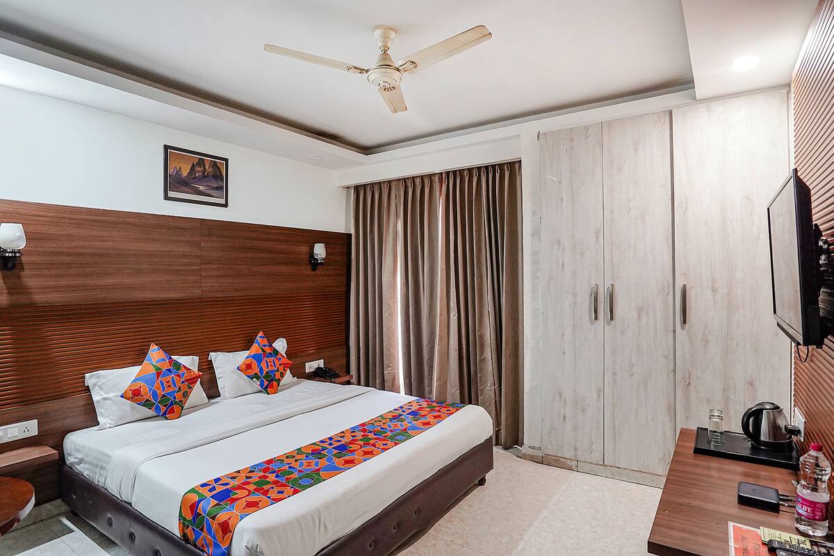 Habitación de hotel con cama, ventilador de techo, televisor, armario y escritorio con tetera y botella.