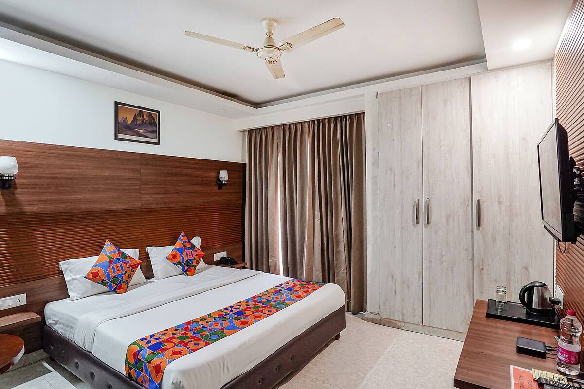 Habitación de hotel con cama, ventilador de techo, televisor, armario y mesita de noche con tetera y botella.