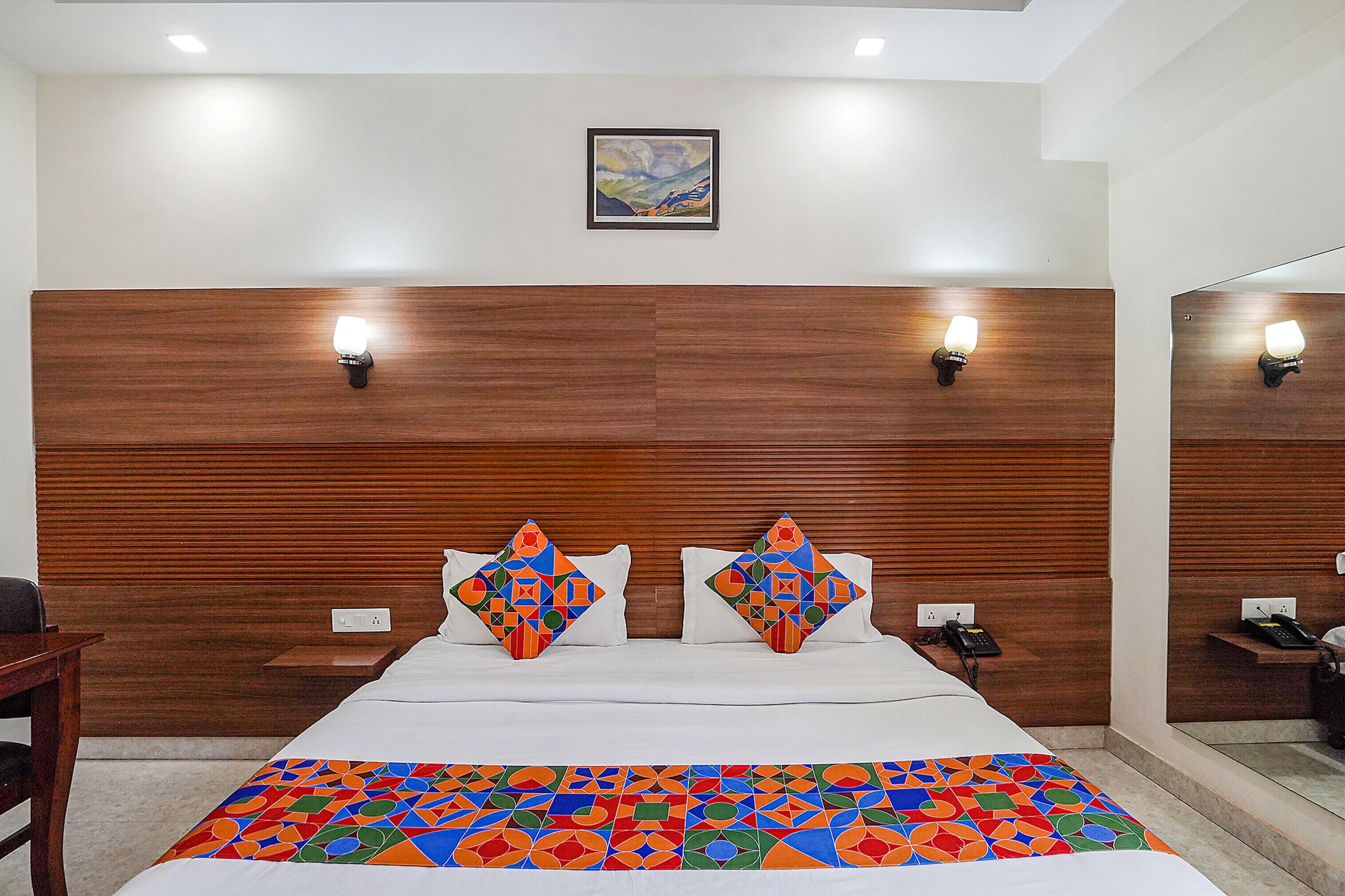 Une chambre d’hôtel avec une tête de lit en bois, deux lampes de chevet, un lit recouvert d’une couvre-lit aux motifs géométriques colorés et un tableau accroché au mur.