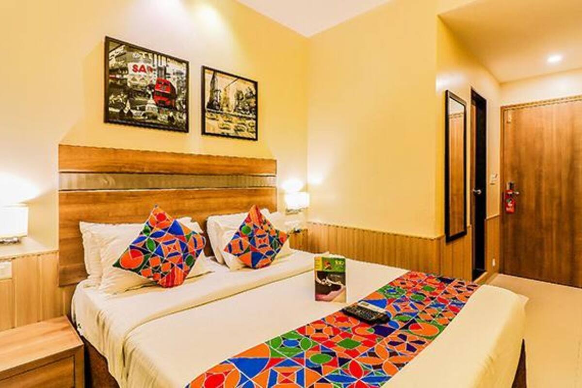 Quarto de hotel com uma cama, duas pinturas na parede, uma porta de madeira e uma mesa de cabeceira com abajur.
