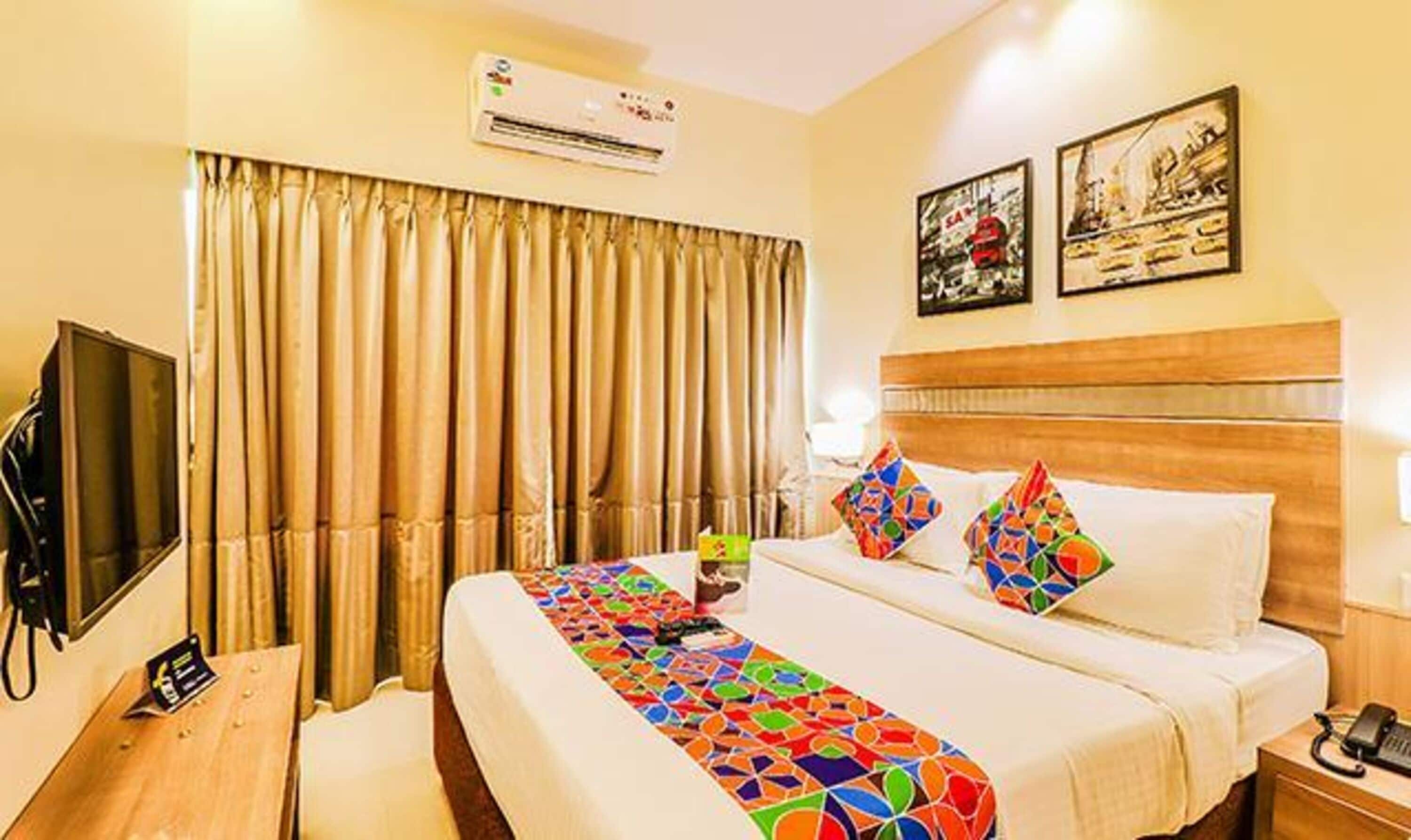 A hotel room with a bed, a desk, an air conditioner, curtains, and two framed pictures on the wall.