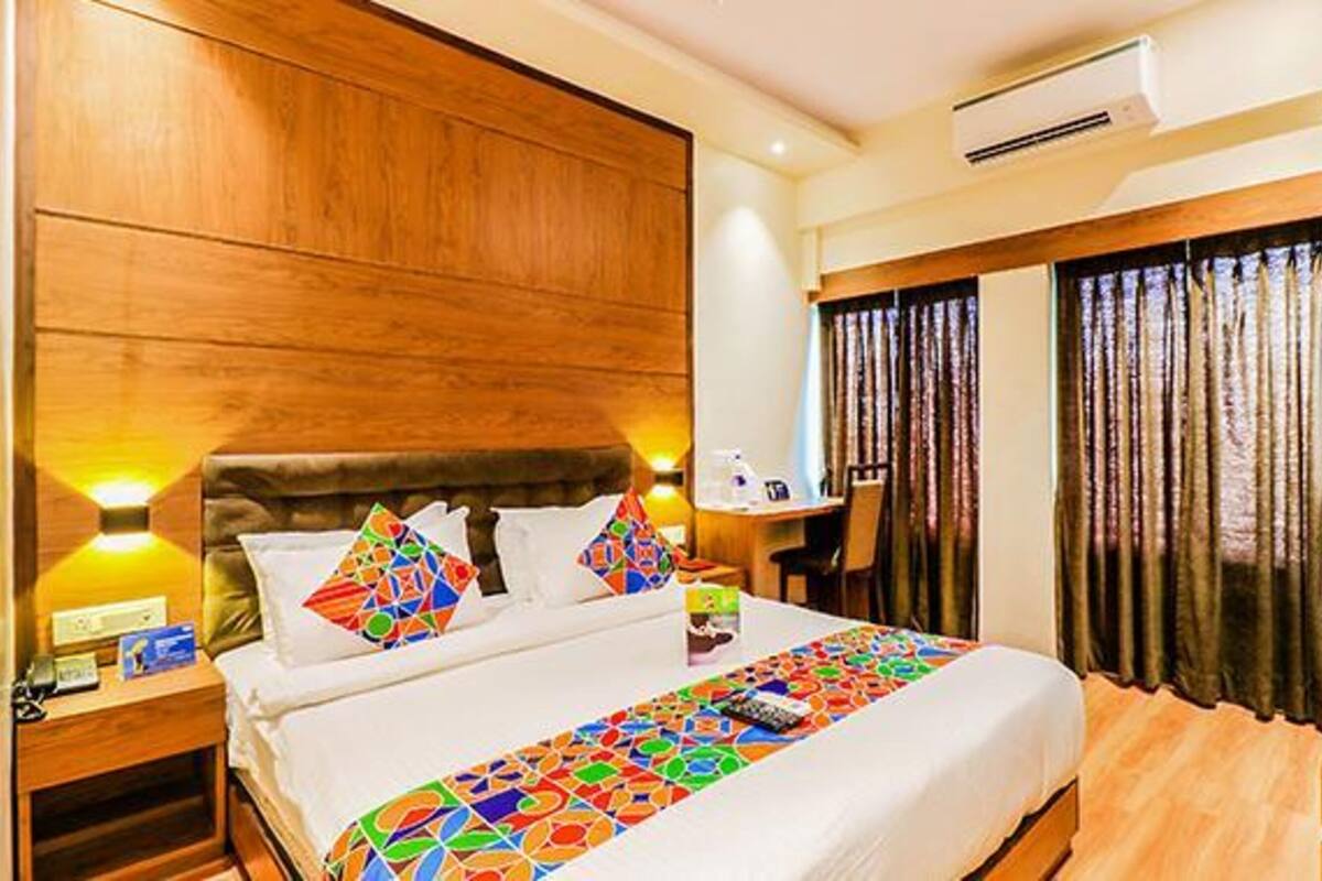 Quarto de hotel com cabeceira de madeira, cama com roupa de cama colorida, mesa de cabeceira com abajur e janela com cortinas.
