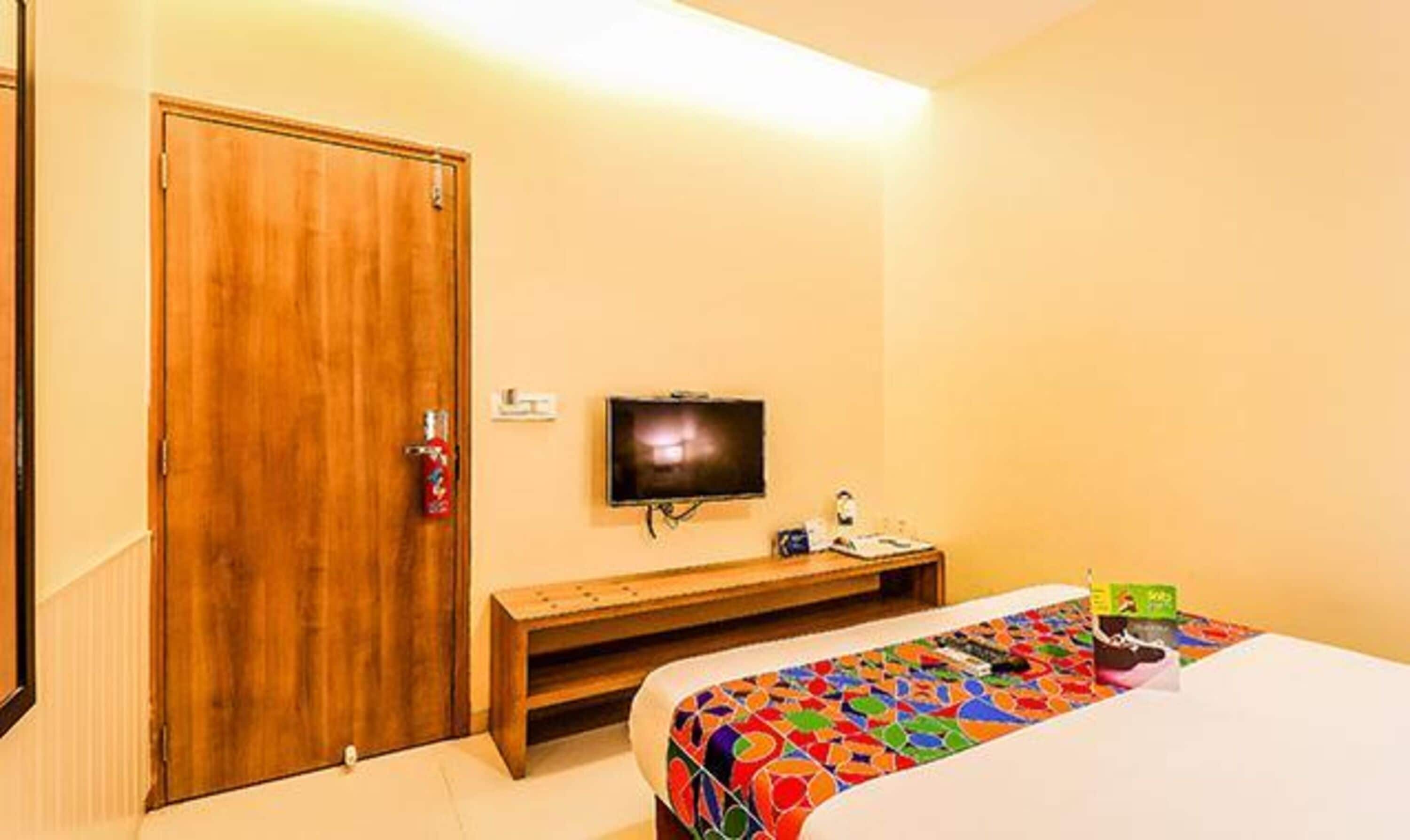 A hotel room with a wooden door, a bed with a colorful bedspread, a television on the wall, and a small table with a lamp and a box.