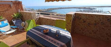 A balcony with a table set for two, a chair, a small potted plant, and a view of the sea and marina.