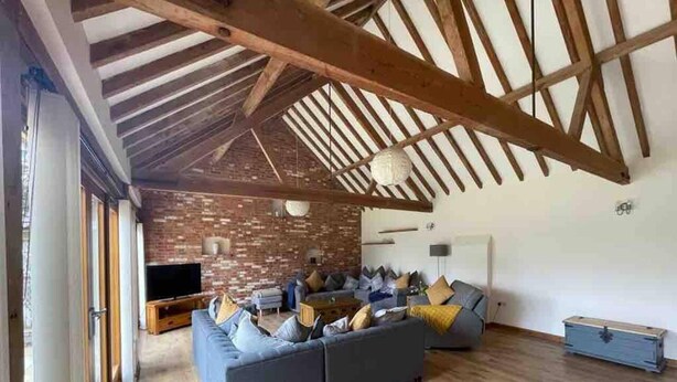 A spacious living room with a large gray sofa, wooden beams on the ceiling, and a brick wall. - Charming Norfolk Barn Conversion in Barnham Broom (Norwich)