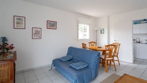 A compact living space with a blue sofa, wooden dining table, and chairs, a kitchenette, and framed pictures on the walls. - Quiet T2 Apartment in Arcachon with Private Parking, Terrace, and Close to Beach and Shops (Arcachon)