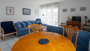 A living room with a blue sofa, a wooden dining table, and a TV on a wooden cabinet. - Quiet T2 Apartment in Arcachon with Private Parking, Terrace, and Close to Beach and Shops (Arcachon)