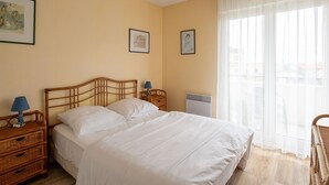 A bedroom with a wooden bed, two bedside tables with lamps, a radiator, and two framed pictures on the wall. - Quiet T2 Apartment in Arcachon with Private Parking, Terrace, and Close to Beach and Shops (Arcachon)
