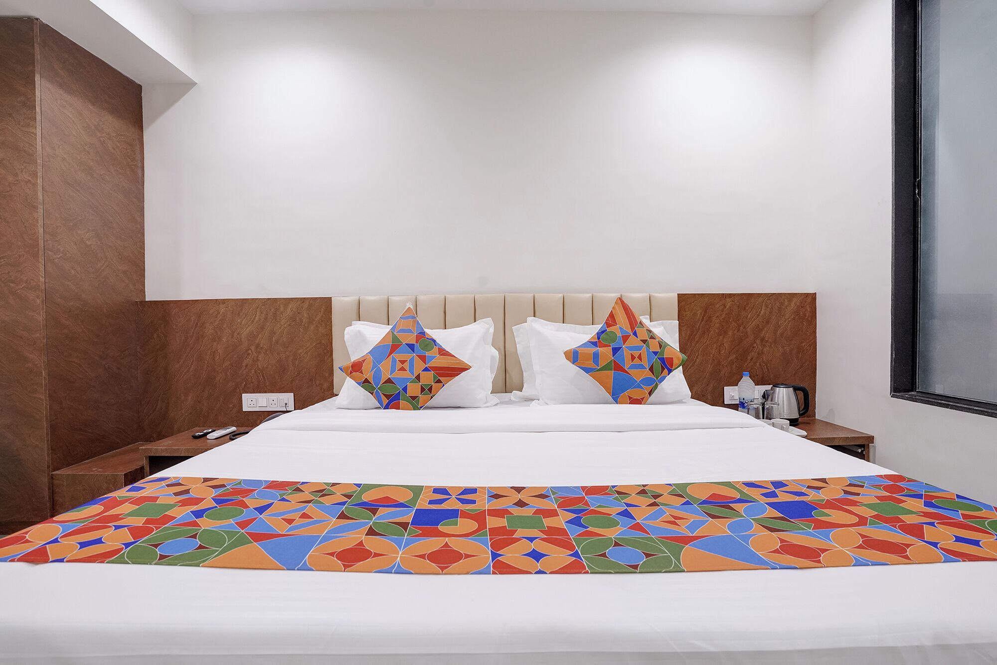 A hotel room with a bed, two pillows, a nightstand, a water bottle, a kettle, and a window.