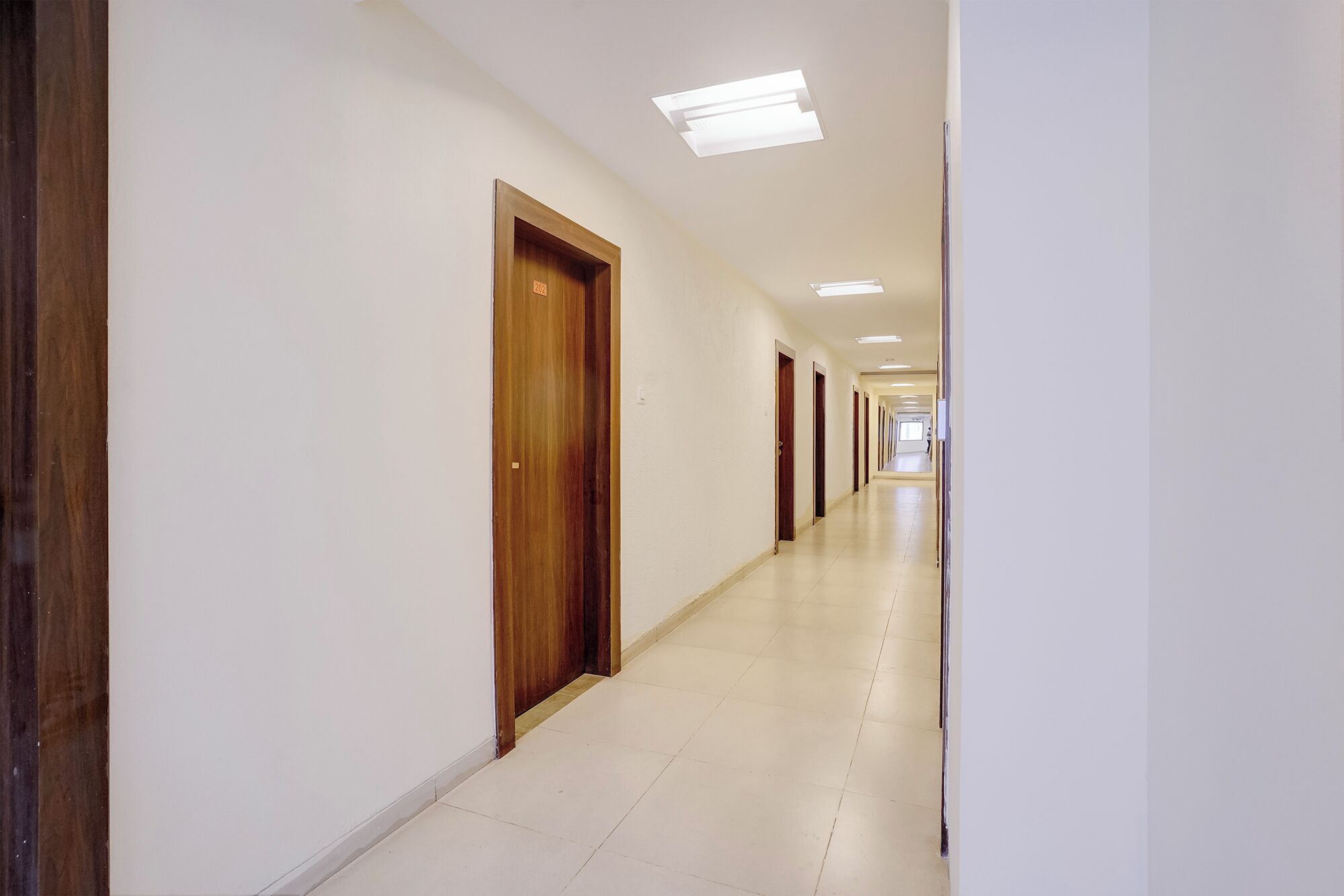 Un largo y estrecho pasillo con puertas de madera y paredes blancas, que conduce a una habitación luminosa al final.