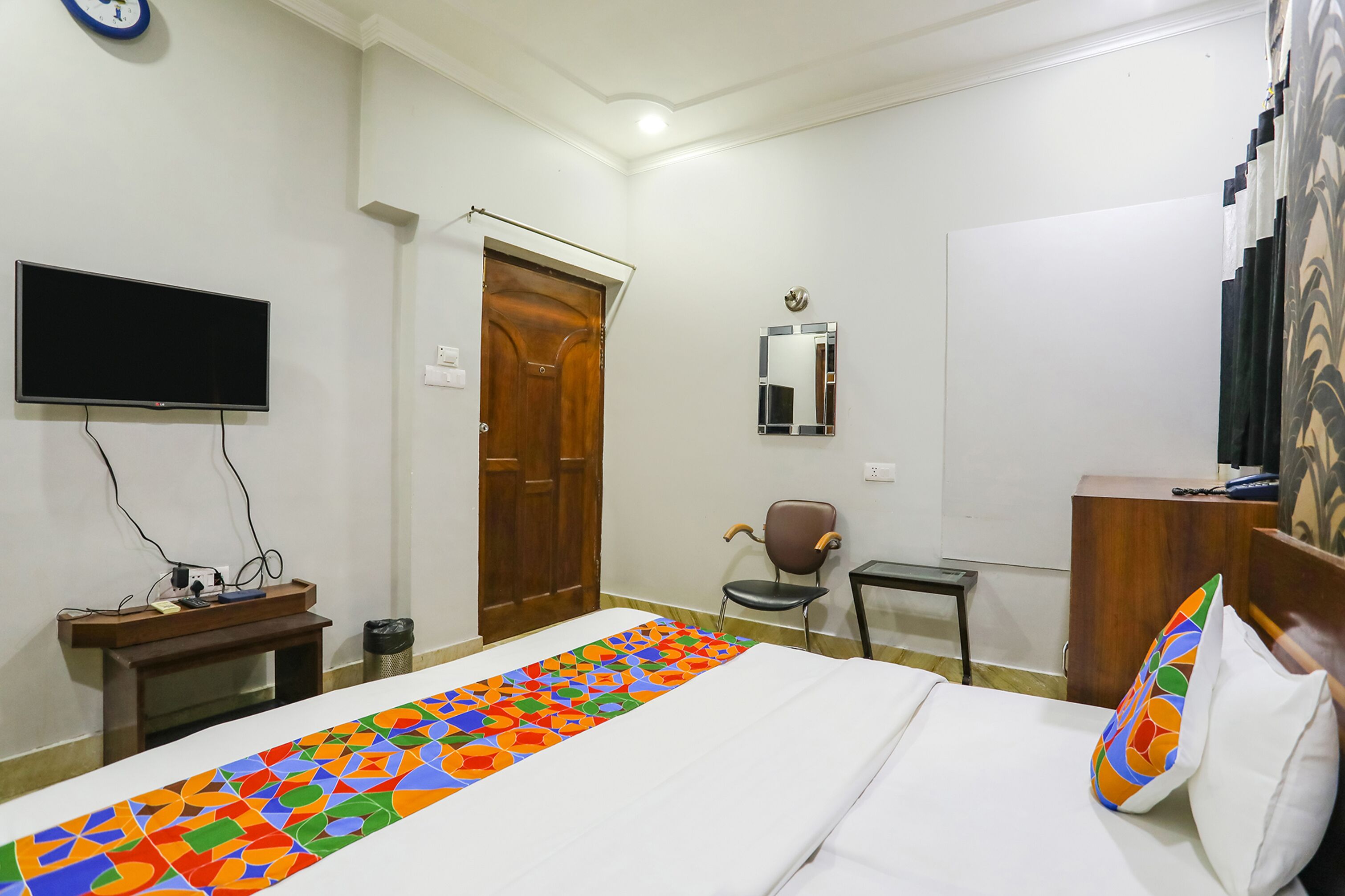 A hotel room with a bed, a TV mounted on the wall, a wooden door, a chair, a small table, and a mirror on the wall.