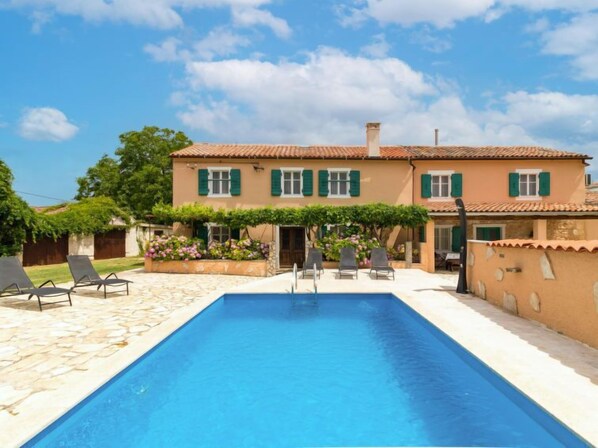 Una casa de dos pisos con piscina, contraventanas verdes y mobiliario de exterior.