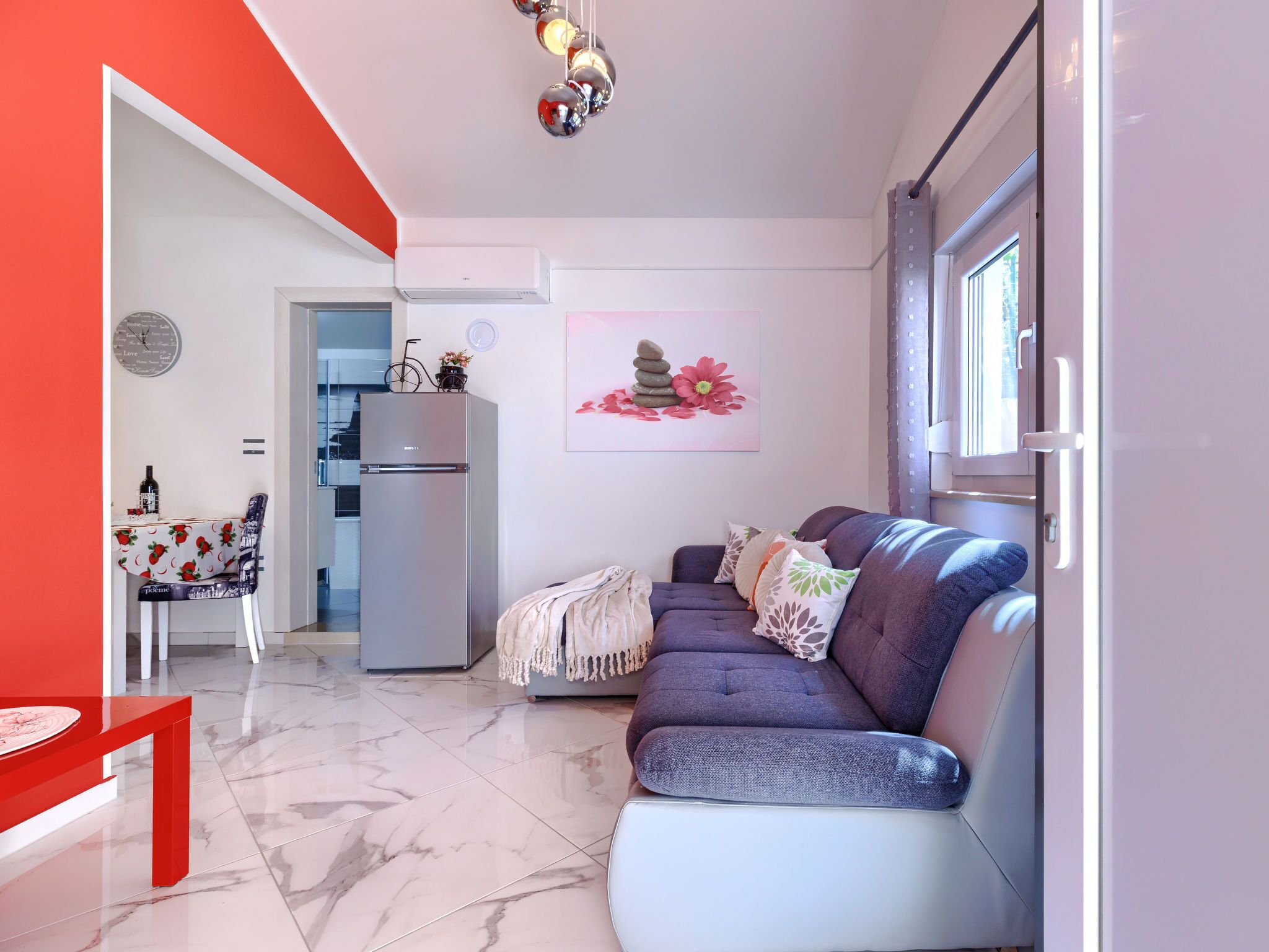 A modern living room with a sofa, a dining table with chairs, a refrigerator, and a kitchen area.