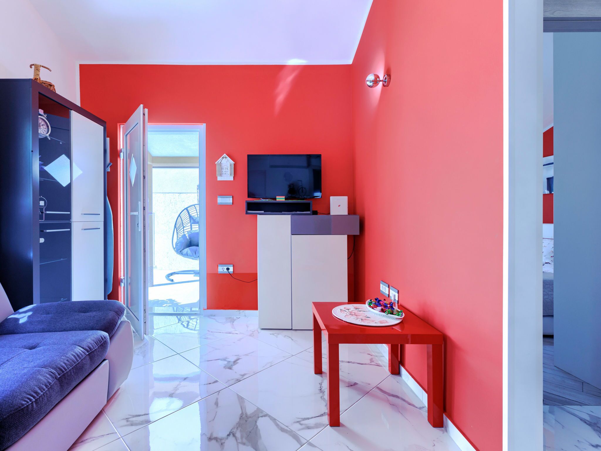 A modern room with a red accent wall, a TV mounted above a white cabinet, a small table with snacks, and a blue sofa.