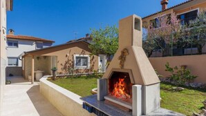 An outdoor stone fireplace with a fire, surrounded by a concrete base and steps, in a residential setting with a clear sky. - Dragica by Interhome (Pula)