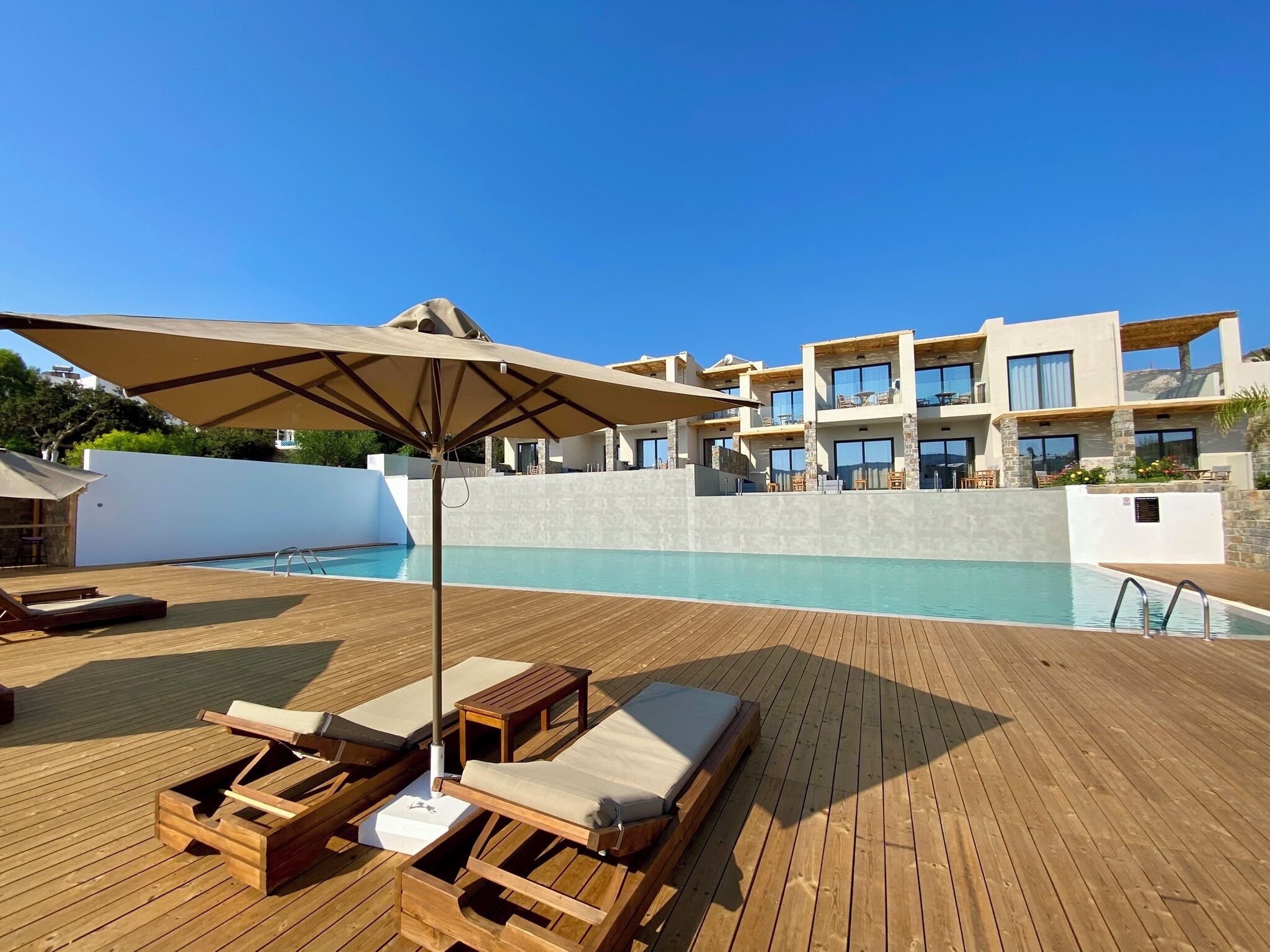 Una terraza de madera con sillones de descanso y una sombrilla, una piscina y un edificio con varios balcones al fondo.