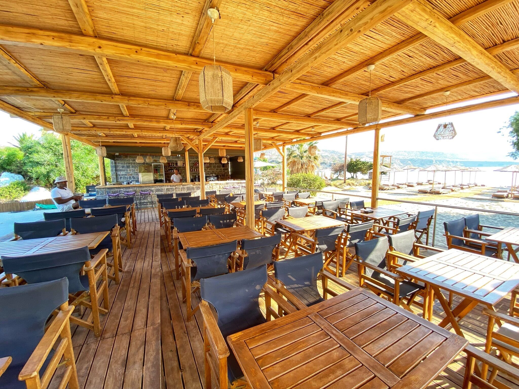 Una terraza de madera con mesas y sillas, una zona de bar y vistas al mar.
