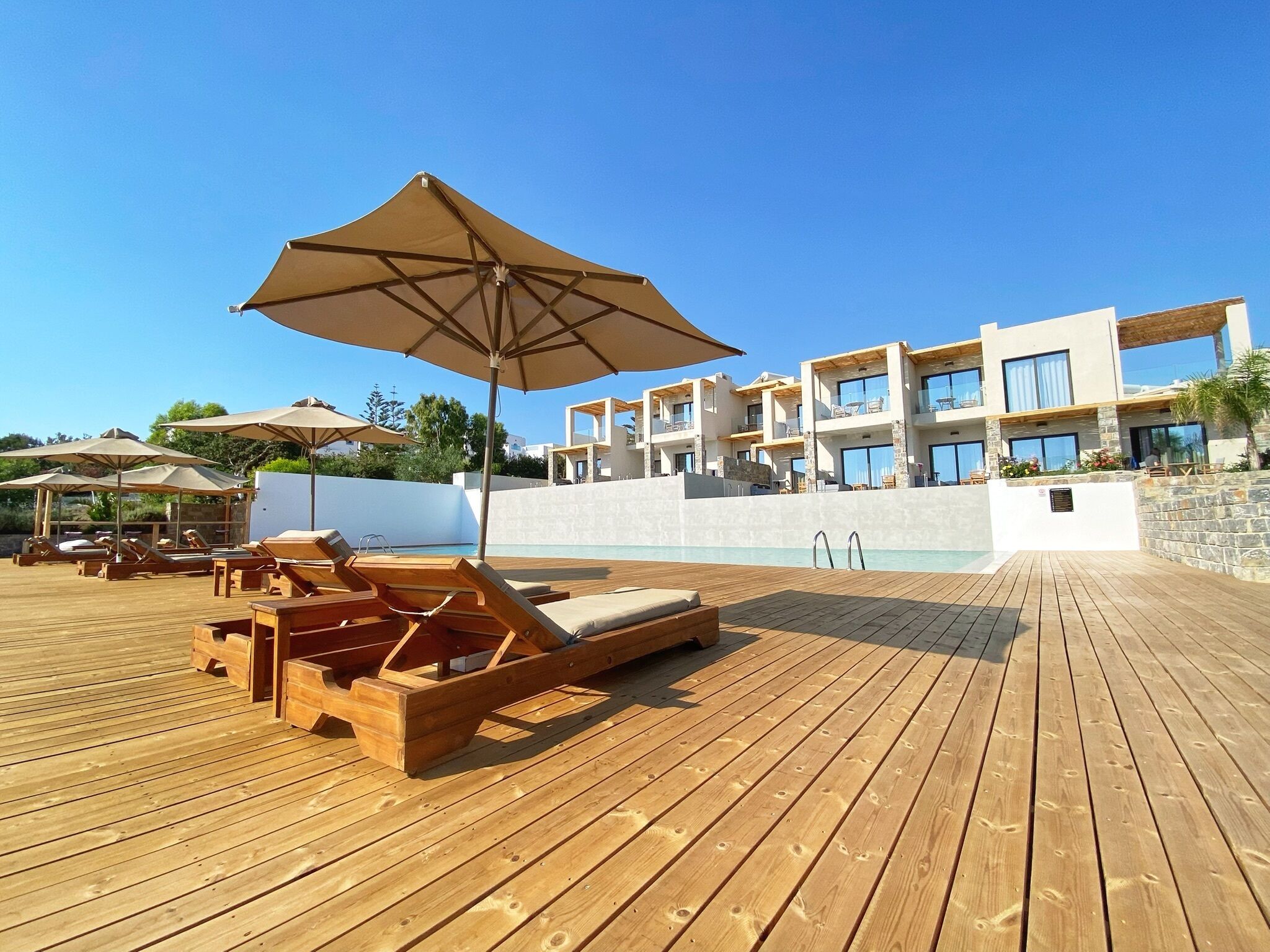 En træterrasse med liggestole og parasoller, en swimmingpool og en moderne bygning med balkoner.