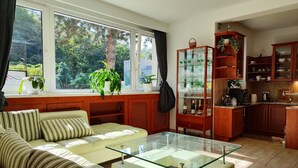 Een moderne woonkamer met een salontafel met glazen blad, een gestreept bankje en uitzicht op een boom door grote ramen.