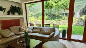 Een moderne woonkamer met een beigekleurige leren bank, een glazen salontafel en een rond hocker. Er is een groot raam met uitzicht op een tuin met bomen en een stenen muur.