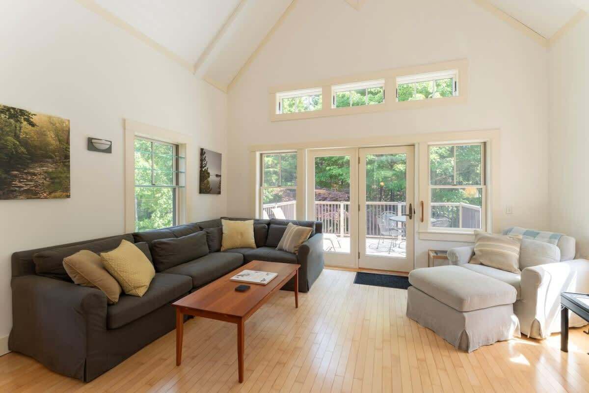 Ein geräumiges Wohnzimmer mit einem dunkelbraunen Ecksofa, einem Holztisch und einem beige-farbenen Hocker. Große Fenster sorgen für Tageslicht und Blick ins Freie.
