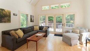 A spacious living room with a dark brown sectional sofa, a wooden coffee table, and a beige ottoman. Large windows provide natural light and a view of the outdoors. - Wonderland Home | 2BR retreat, mins to Acadia NP (Tremont)