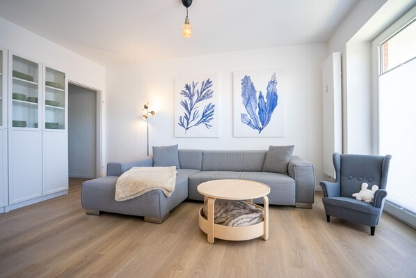 A modern living room with a grey sofa, a round wooden coffee table, a grey armchair, and two wall-mounted light fixtures. - Herzlich Willkommen in Unserem Liebevoll Eingerichtetem Appartement "albatros" (Büsum)