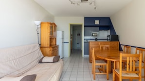 A living room with a sofa, wooden cabinet, and a dining table with chairs. A kitchen is visible in the background. - Mimizan Beach: 6-person apartment with balcony and parking (Mimizan)