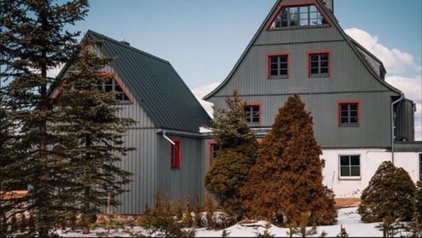 Une maison à deux étages, avec un toit gris et des encadrements de fenêtres rouges, entourée d’arbres et de neige.