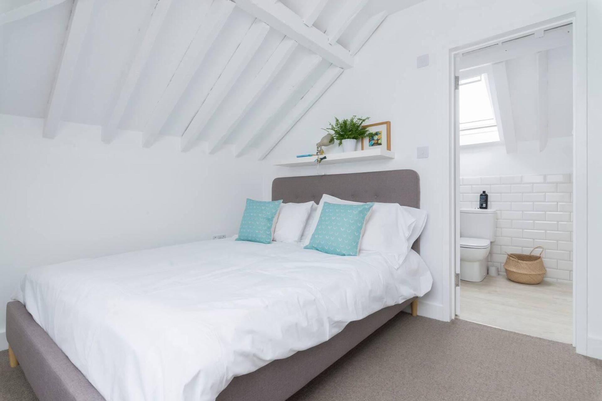 A bedroom with a bed, a grey headboard, white walls, a white ceiling, a small shelf with a plant, and a view into a bathroom with a toilet and a basket.