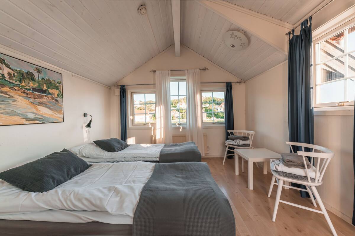 A room with two beds, a small table, and a chair. There are windows with curtains, a painting on the wall, and a ceiling light.