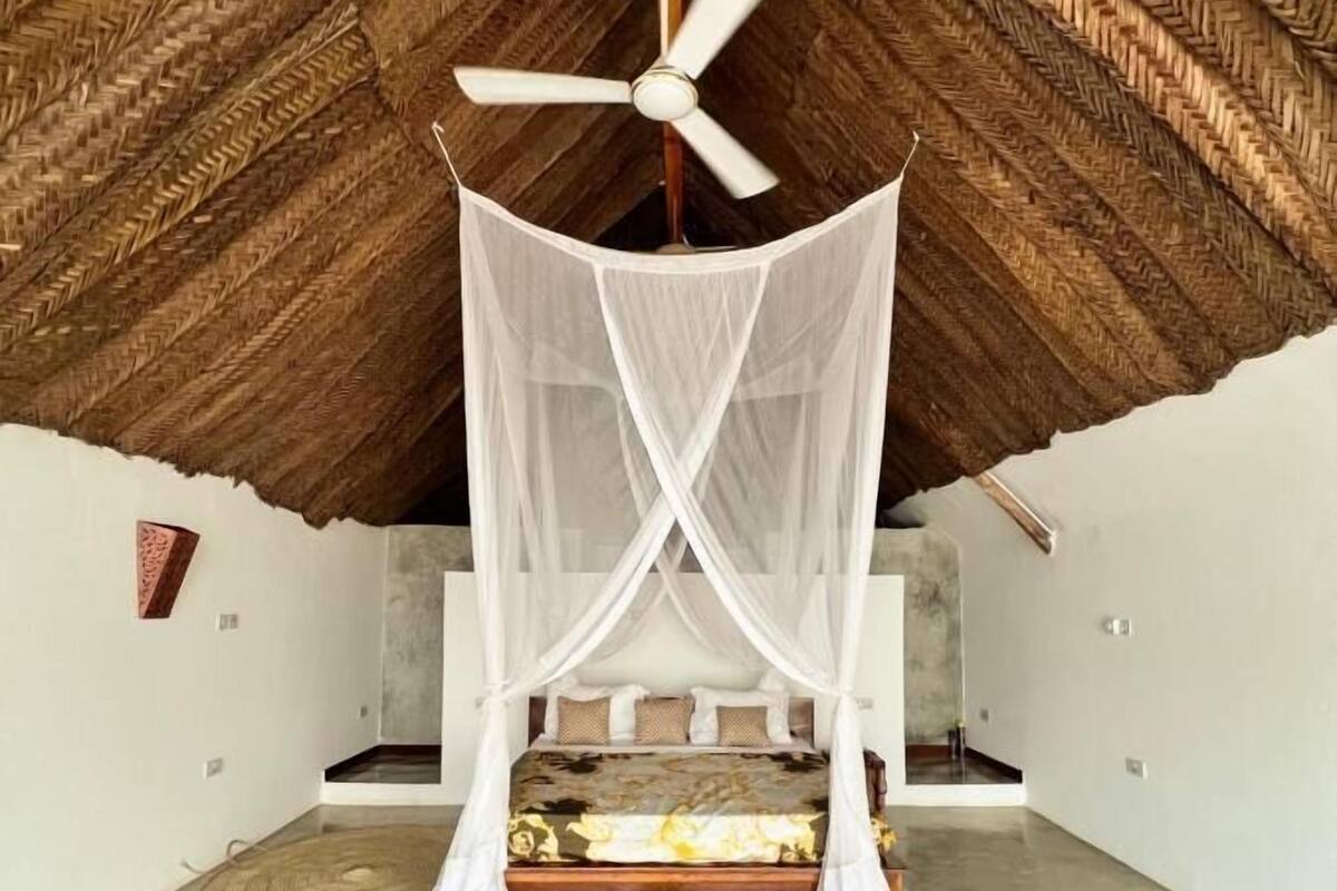 A room with a thatched roof, a bed with a canopy, and a ceiling fan.