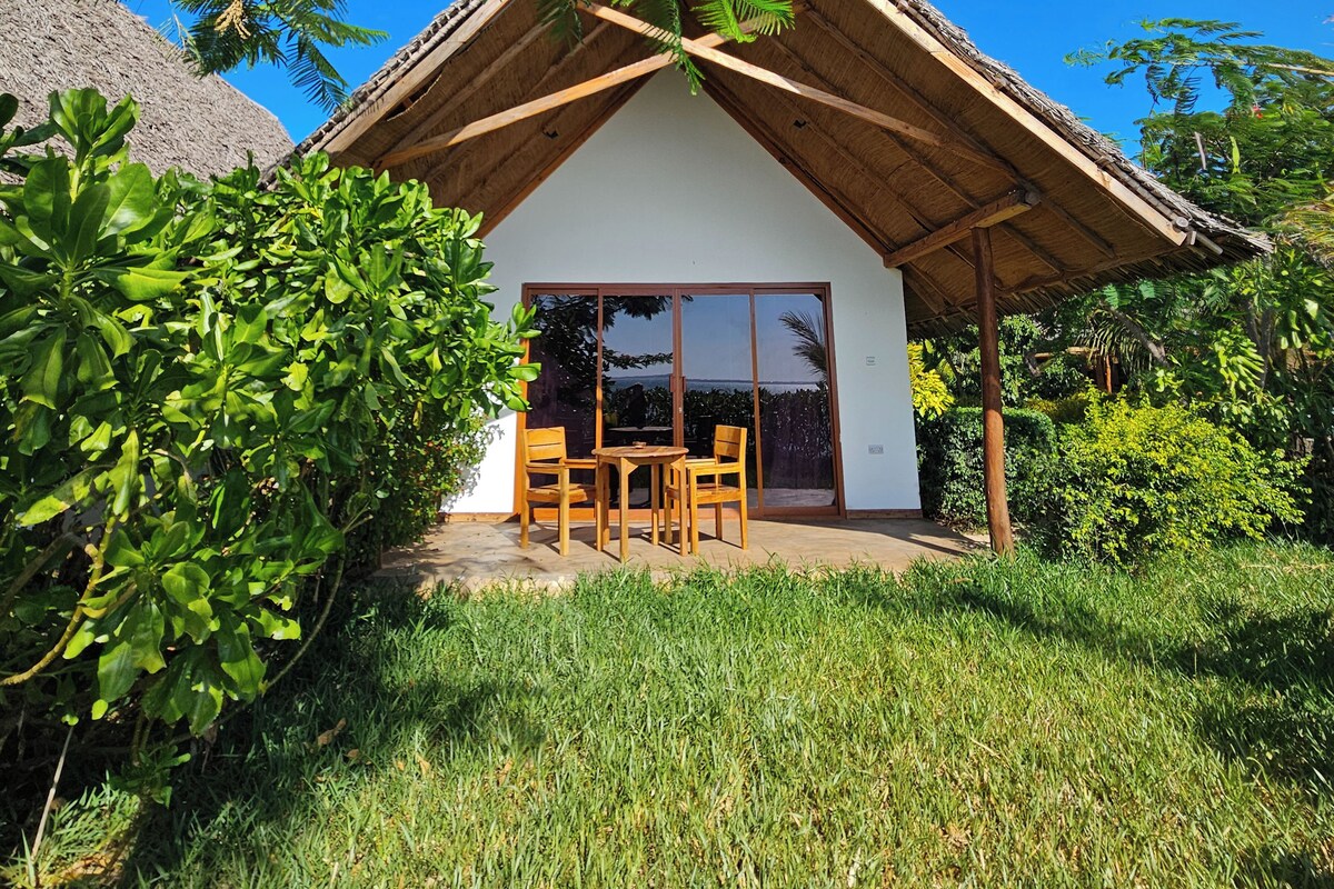 A house with a thatched roof, a covered patio with wooden furniture, and a view of the surrounding greenery.