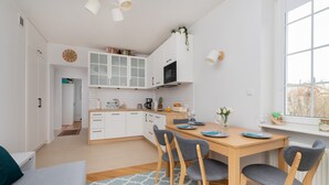 Una cocina moderna con armarios blancos, una mesa de comedor de madera y sillas. Hay un reloj en la pared y una ventana con vistas a la ciudad.