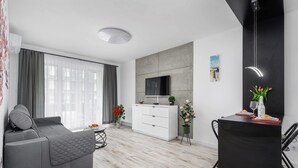 Un salon moderne avec un canapé gris, un meuble blanc, une table à manger recouverte d’une nappe noire et un téléviseur fixé à un mur gris.