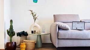 Una sala moderna con un sofá gris, una mesita redonda pequeña, un cactus, un jarrón con plantas secas y un ordenador portátil sobre el sofá.