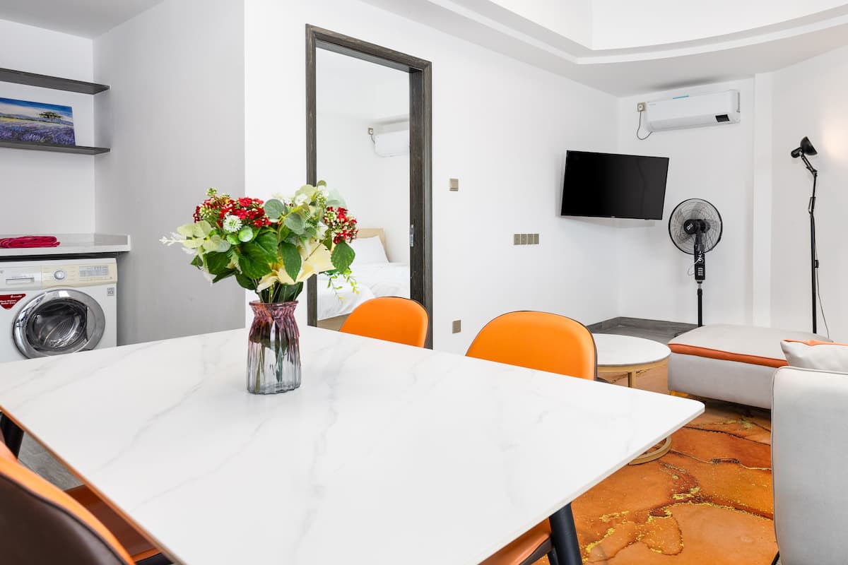 Ein modernes Zimmer mit einem Marmortisch, orangefarbenen Stühlen, einem weißen Sofa, einem Fernseher, einem Ventilator und einer Waschmaschine.