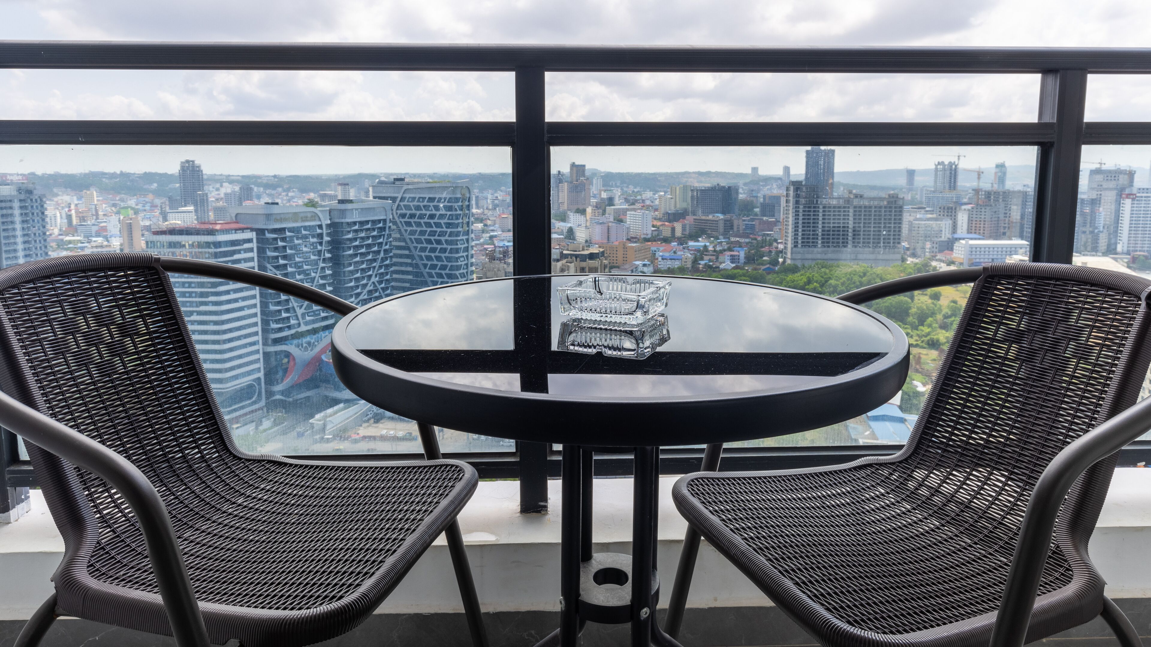 A round table with two wicker chairs, a glass ashtray, and a cityscape view through large windows.