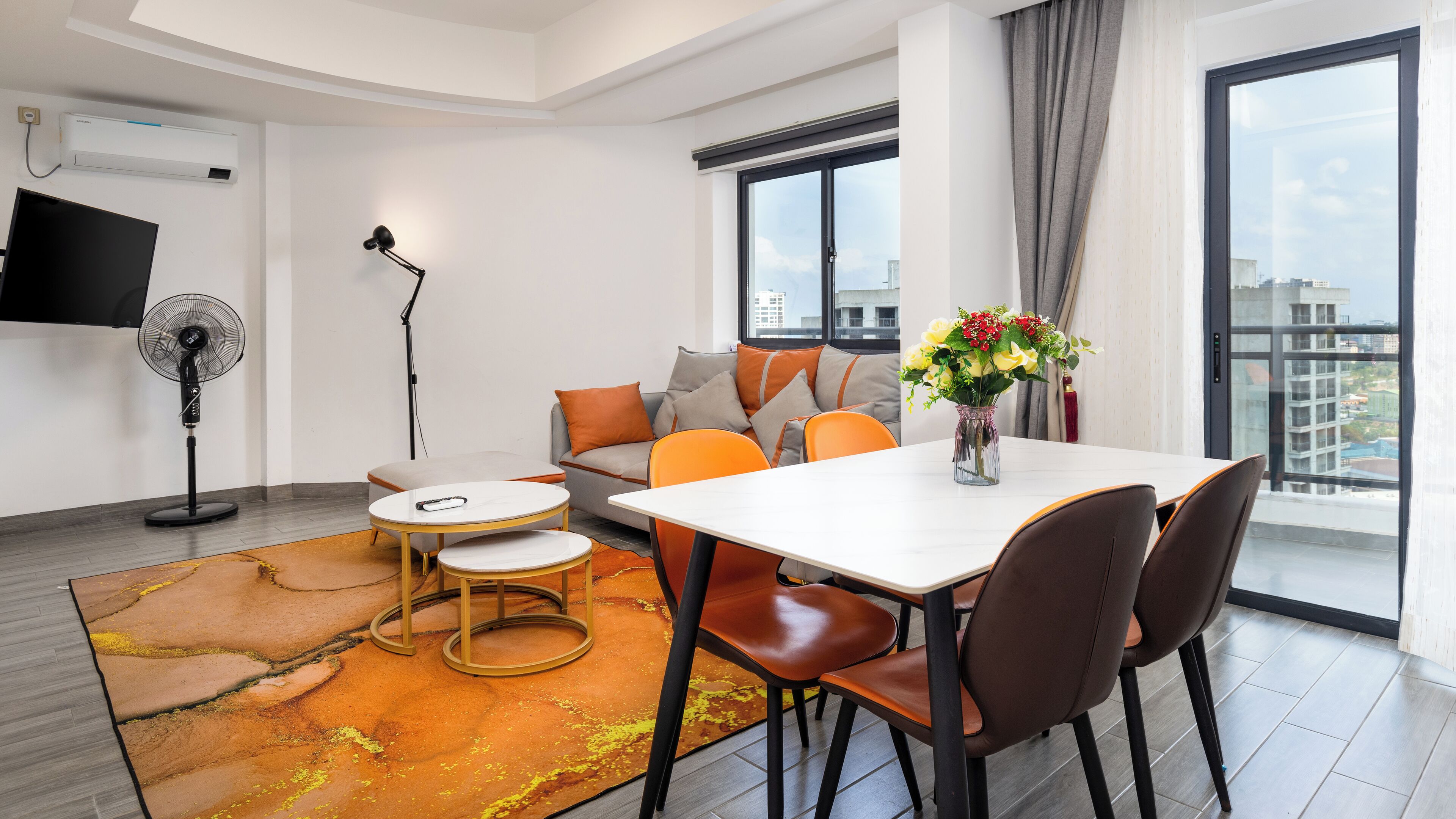 A modern living room with a sofa, a dining table with chairs, a standing fan, and a TV mounted on the wall.