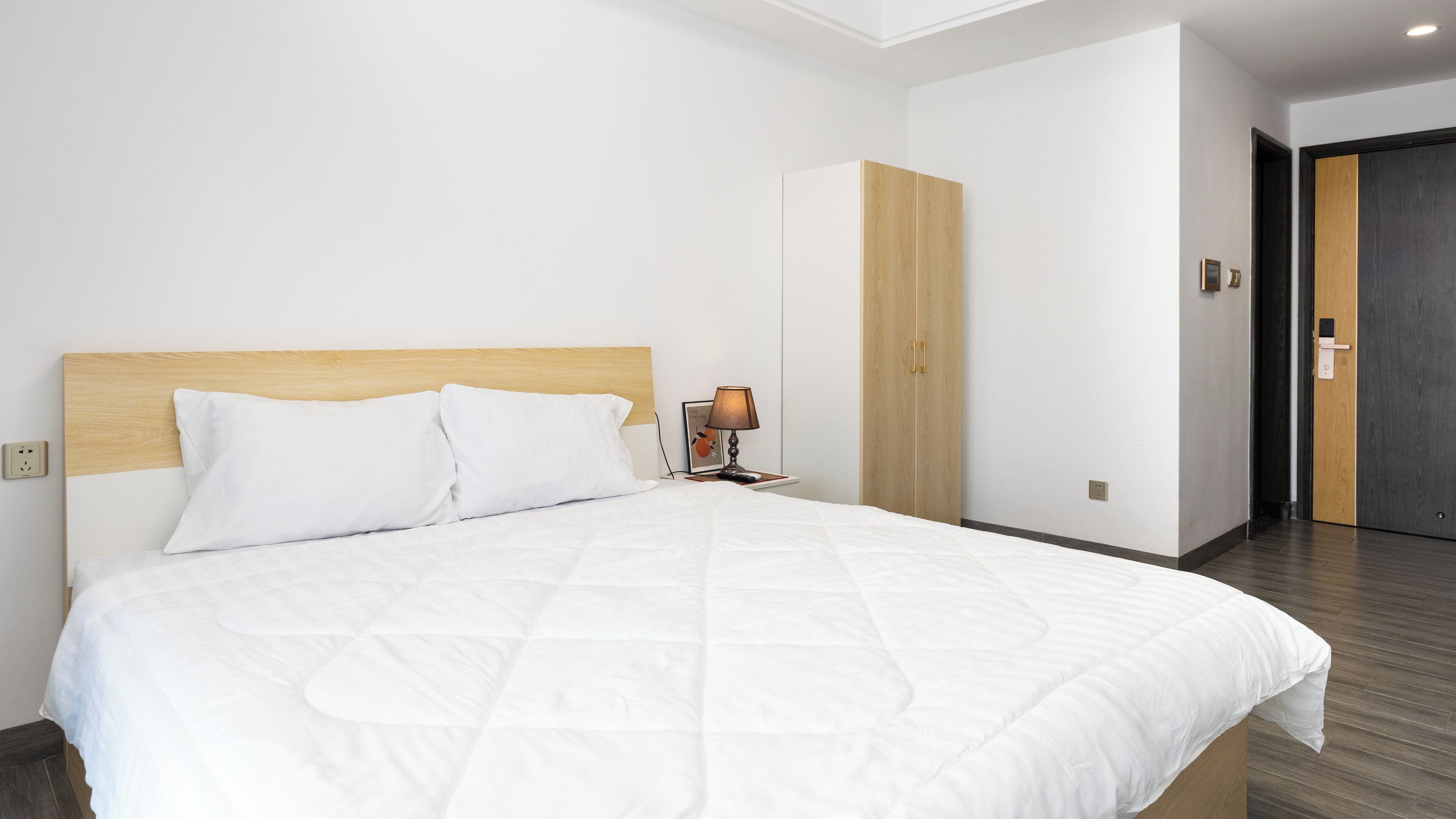 Quarto de hotel com cama grande, mesa de cabeceira com abajur, guarda-roupa e uma porta.