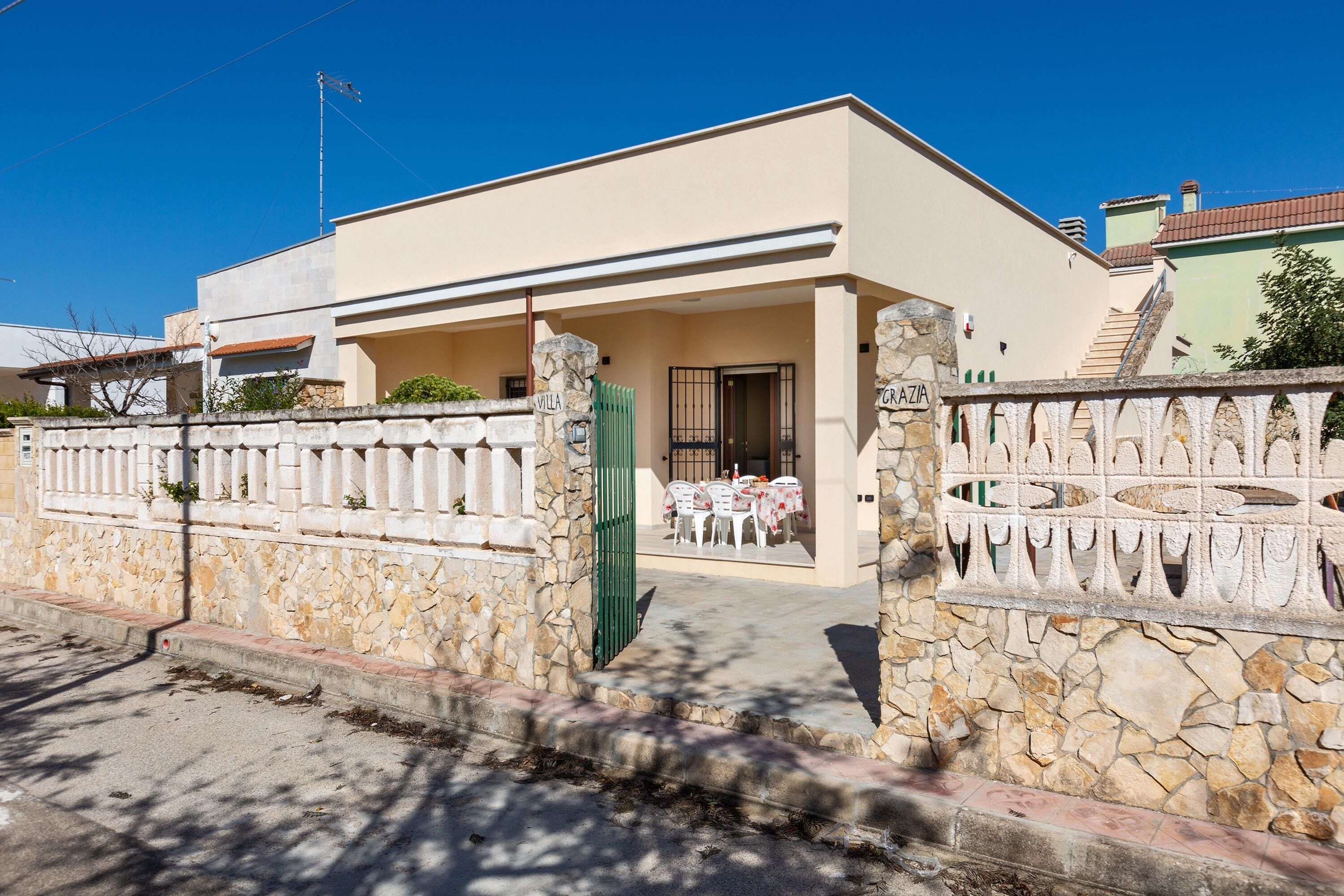 Une maison à deux étages avec une entrée surmontée d’un mur en pierre, un patio couvert avec des meubles d’extérieur et un ciel d’un bleu éclatant.