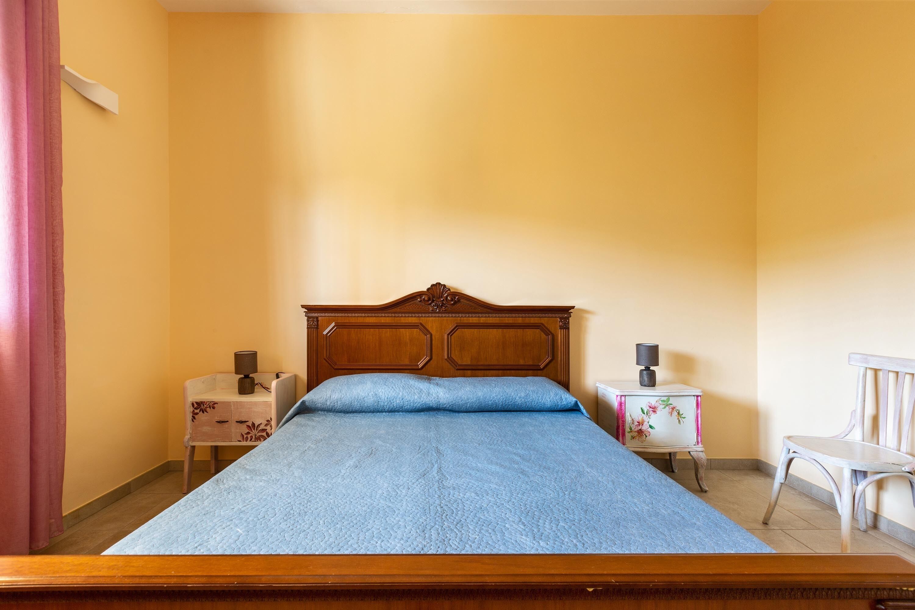 A bedroom with a wooden bed, a nightstand, a chair, and a small table with a lamp.