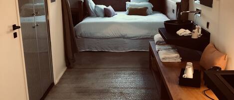 Habitación de hotel con cama, mesita de noche con espejo, lavamanos pequeño y puerta con cortinas.