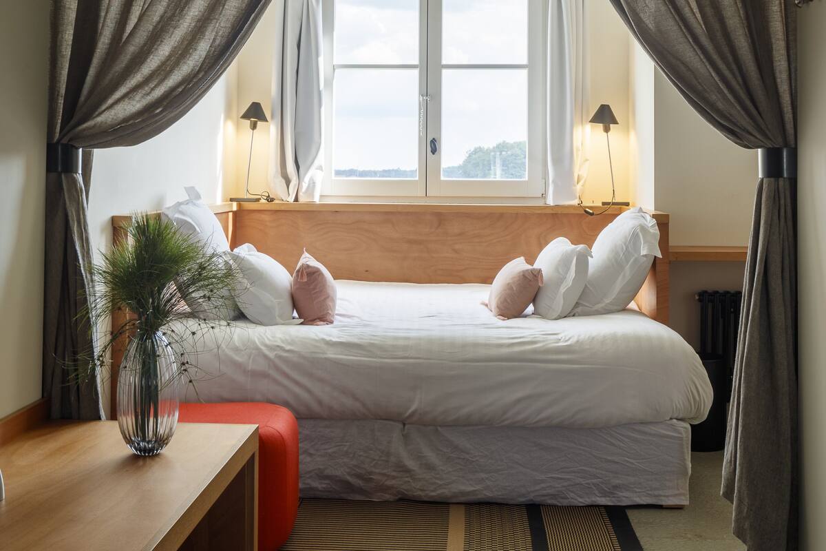 A bedroom with a bed, a bedside table, a lamp, a window, and curtains.