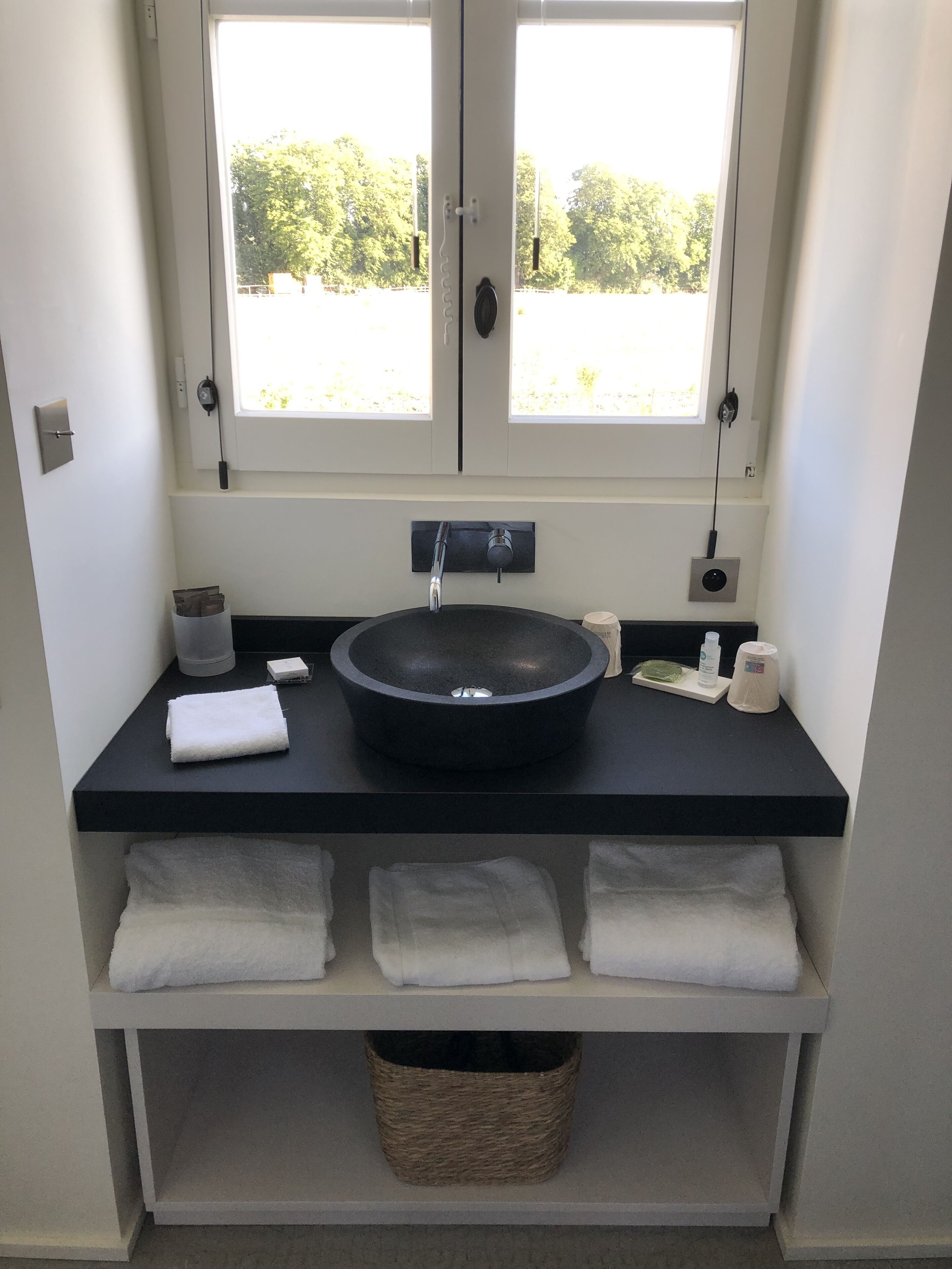 A bathroom with a black sink, two towel racks, a small basket, and a window with a view of trees.