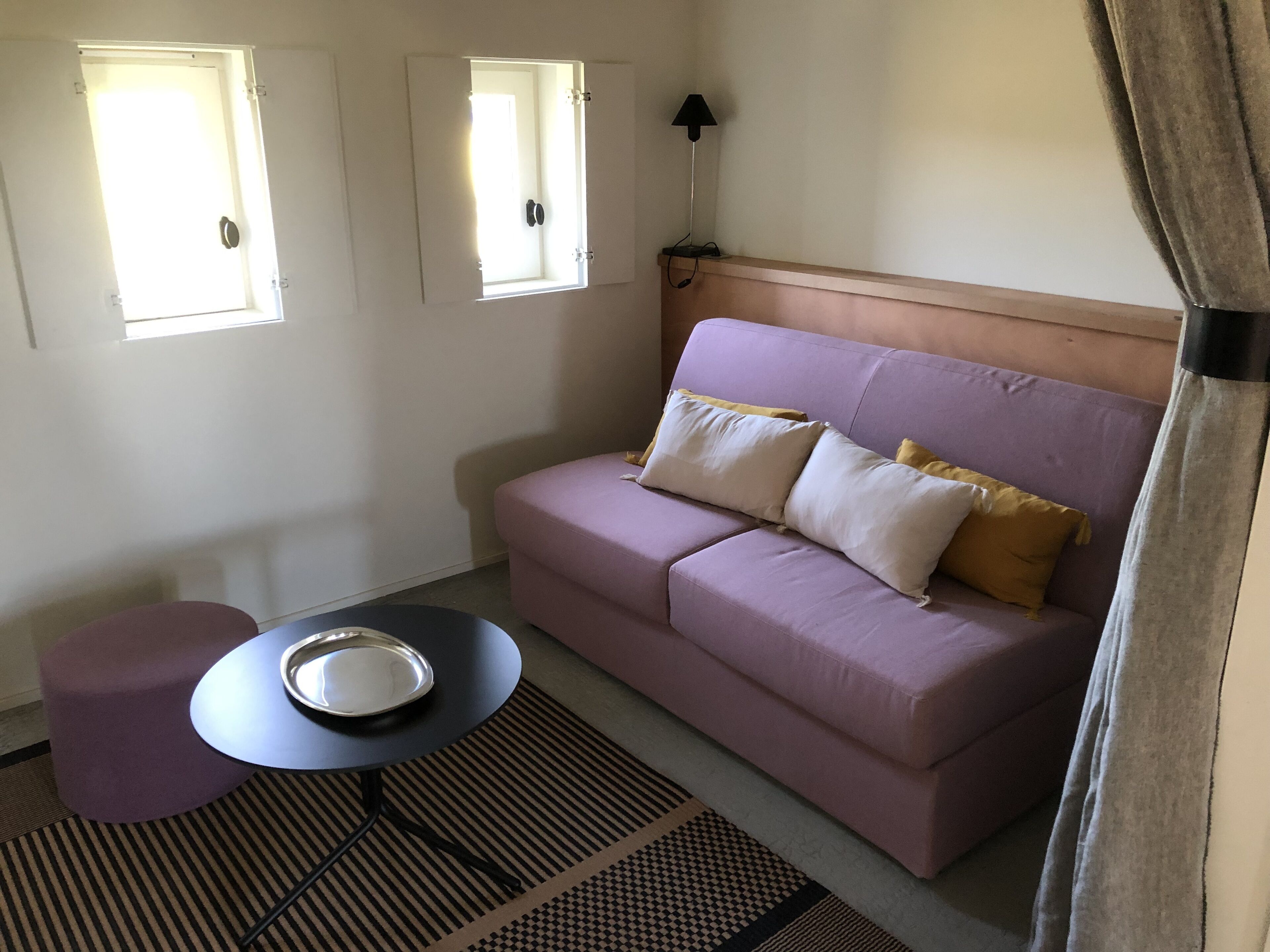 A room with a purple sofa, a small round table with a metal tray, a patterned rug, and two windows with blinds.
