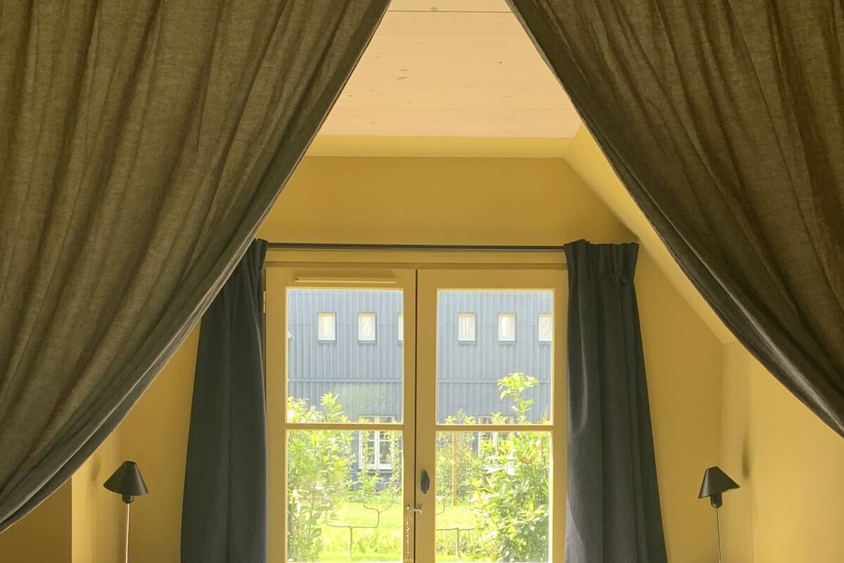 A bedroom with a bed, two pillows, a nightstand, a lamp, and a window with curtains.